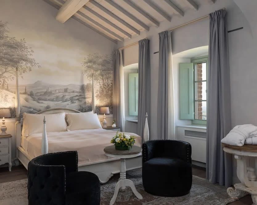 white four poster bed with two black chairs at the food of the bed, grey curtains, and wood beam ceiling with a fresco painted wall